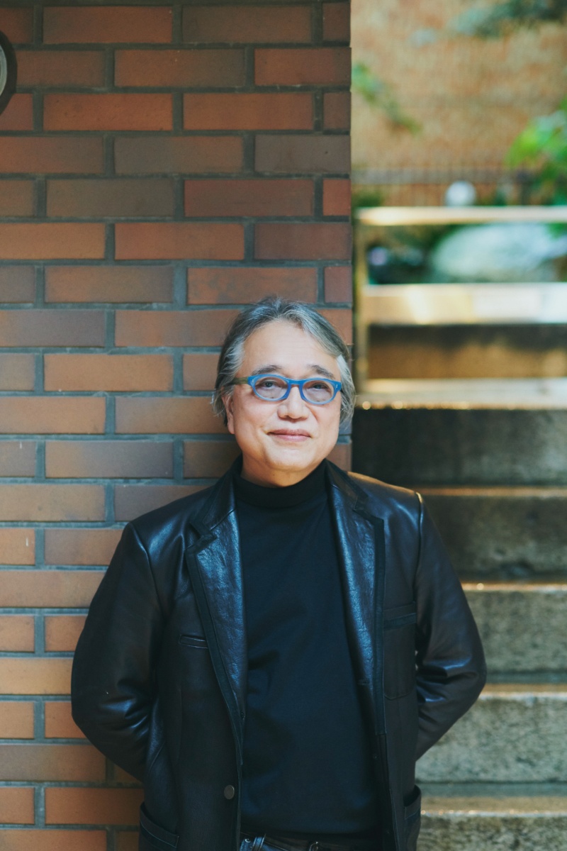 遠藤突無也氏のプロフィール写真。レンガ造りの茶色い壁を背景に立つ男性で、青いフレームの眼鏡をかけ、黒いタートルネックと黒い革ジャケットを着用。背景右奥には階段と観葉植物がぼんやり見える