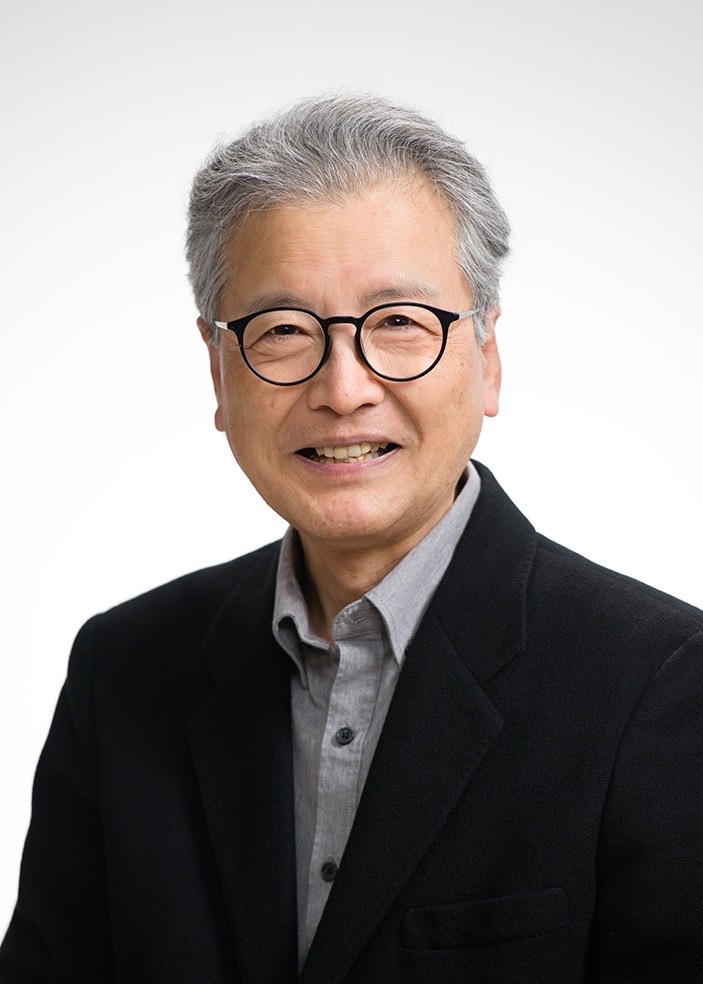 萩原朔美氏のプロフィール写真。白い背景の前に立つ、グレーの短髪と黒い丸フレーム眼鏡をかけた中年男性。グレーのシャツの上に黒いブレザーを着用し、穏やかな笑顔で正面を向いている