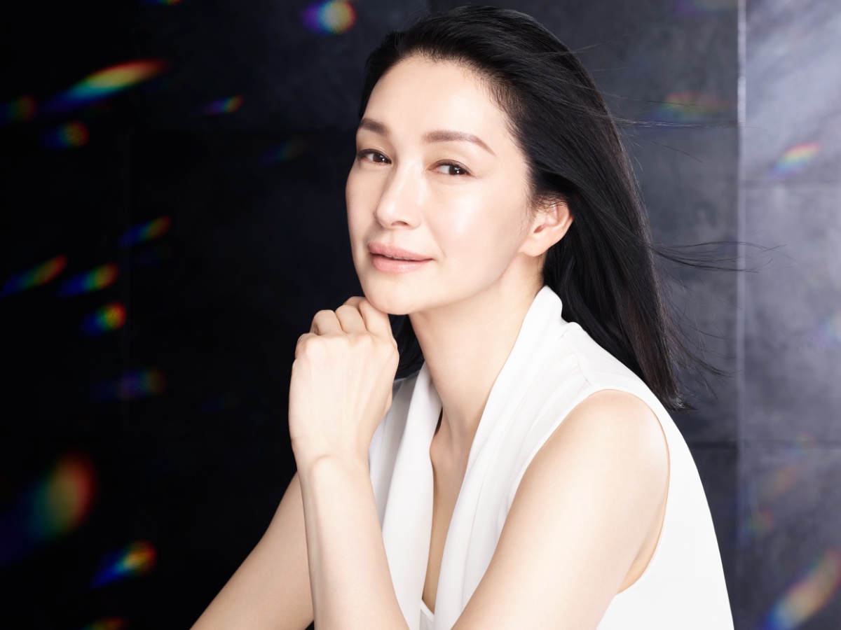 田中久美子氏のプロフィール写真。黒い背景に虹色の光が散在する中、黒い長髪の女性が右向きに立ち、白いノースリーブシャツを着用。落ち着いた表情で肩越しに視線を向けている