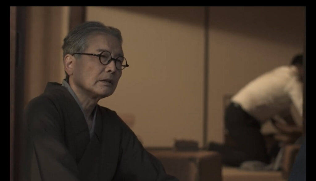 ベージュ色の室内背景で、メガネをかけた中年の日本人男性が黒いシャツを着て、側面を向いて表情豊かに演じている。背景には家具と壁が見える映画や舞台作品のシーン