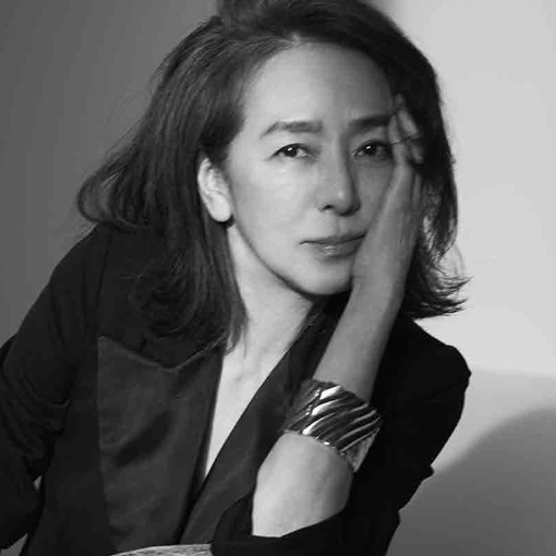 黒いブレザーを着た女性の白黒ポートレート。右手を顔に当てた思慮深い表情で、グレーの背景