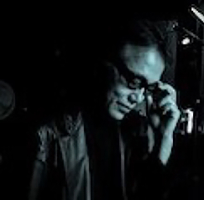 舞台照明の下でマイクを握り演技する人物。暗い背景に浮かび上がるシルエット的な雰囲気で、集中した表情とパフォーマンスの緊迫感が表現されている