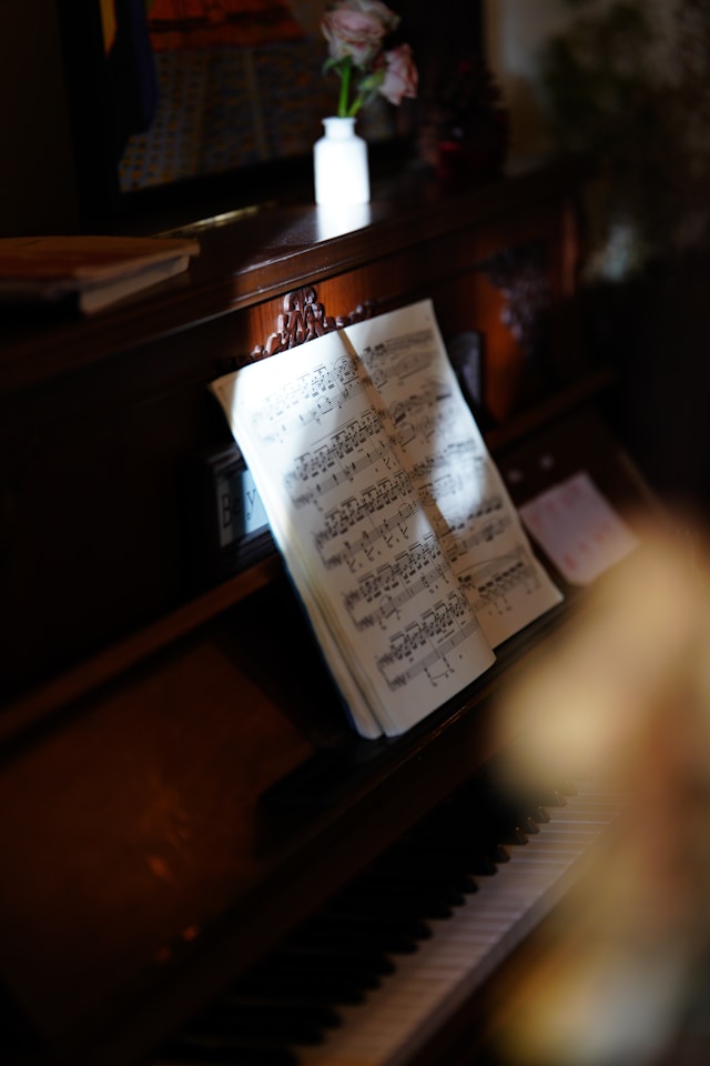 ピアノの上に置かれた白い花瓶と楽譜。暖かい照明に照らされた木製のピアノの鍵盤が手前にぼけて映っている