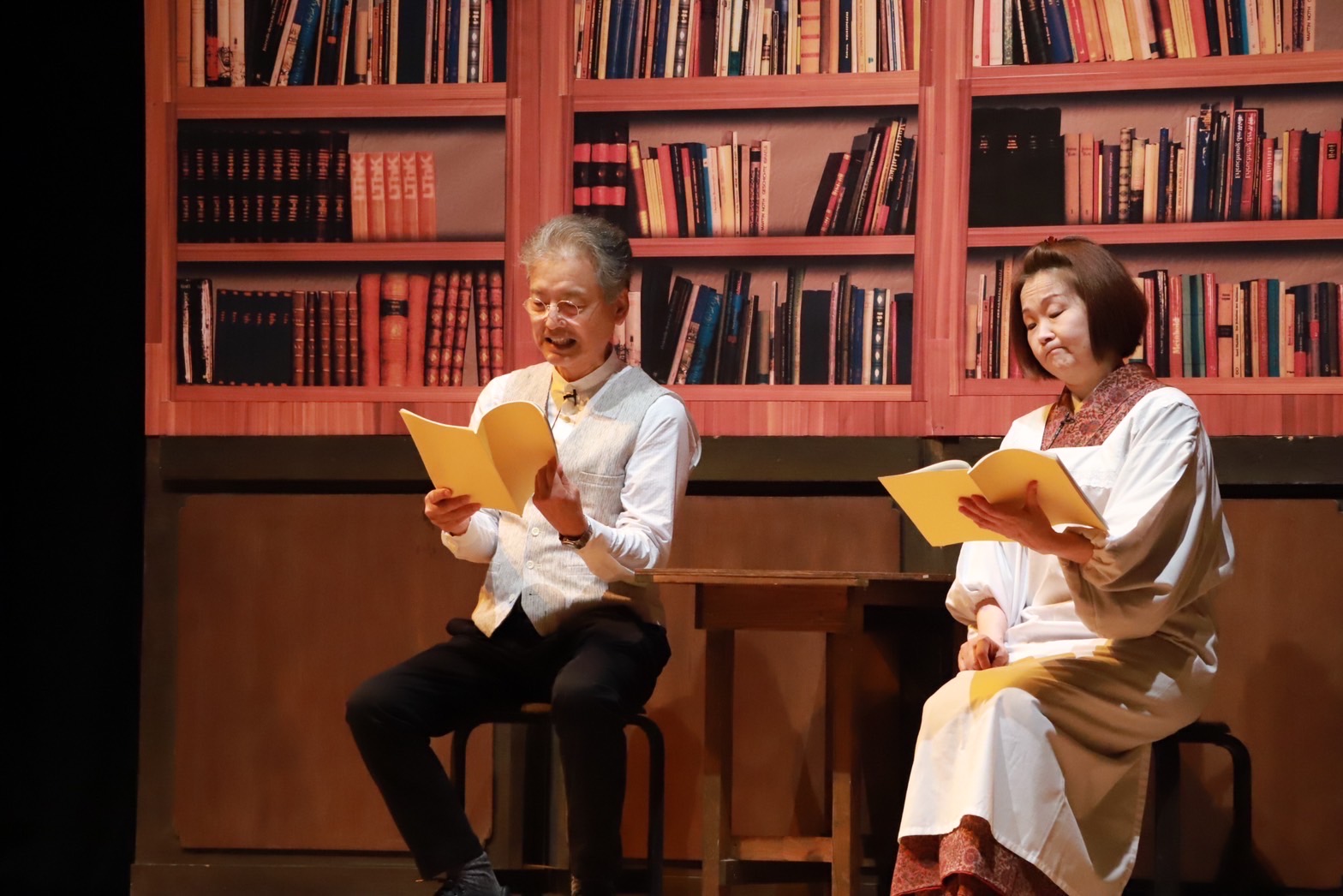 舞台上で赤いシェルフを背景に、白いシャツと黄色の襟のベストを着た日本人中年男性と、白いシャツに紫紋様の羽織を着た日本人女性が、黄色い台本を持ちながら対話している演劇シーン