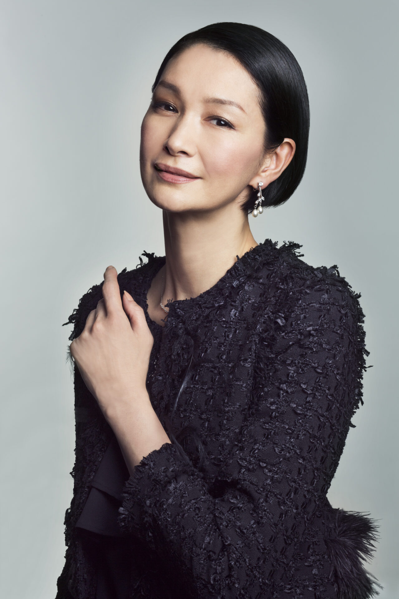 グレーの背景を背に、黒い毛羽立ったテクスチャー素材のトップスを着た日本人女性の胸像。真珠のドロップイヤリングを着用し、上品な表情でカメラを見つめている